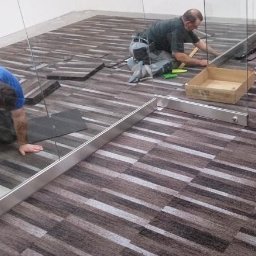 DANIEL ADAMSKI PODŁOGI - Układanie brązowo-szarej wykładziny dywanowej w płytkach. Dwóch monterów pracuje w pomieszczeniu biurowym, widoczne narzędzia pracy i fragment szklanej ścianki działowej.