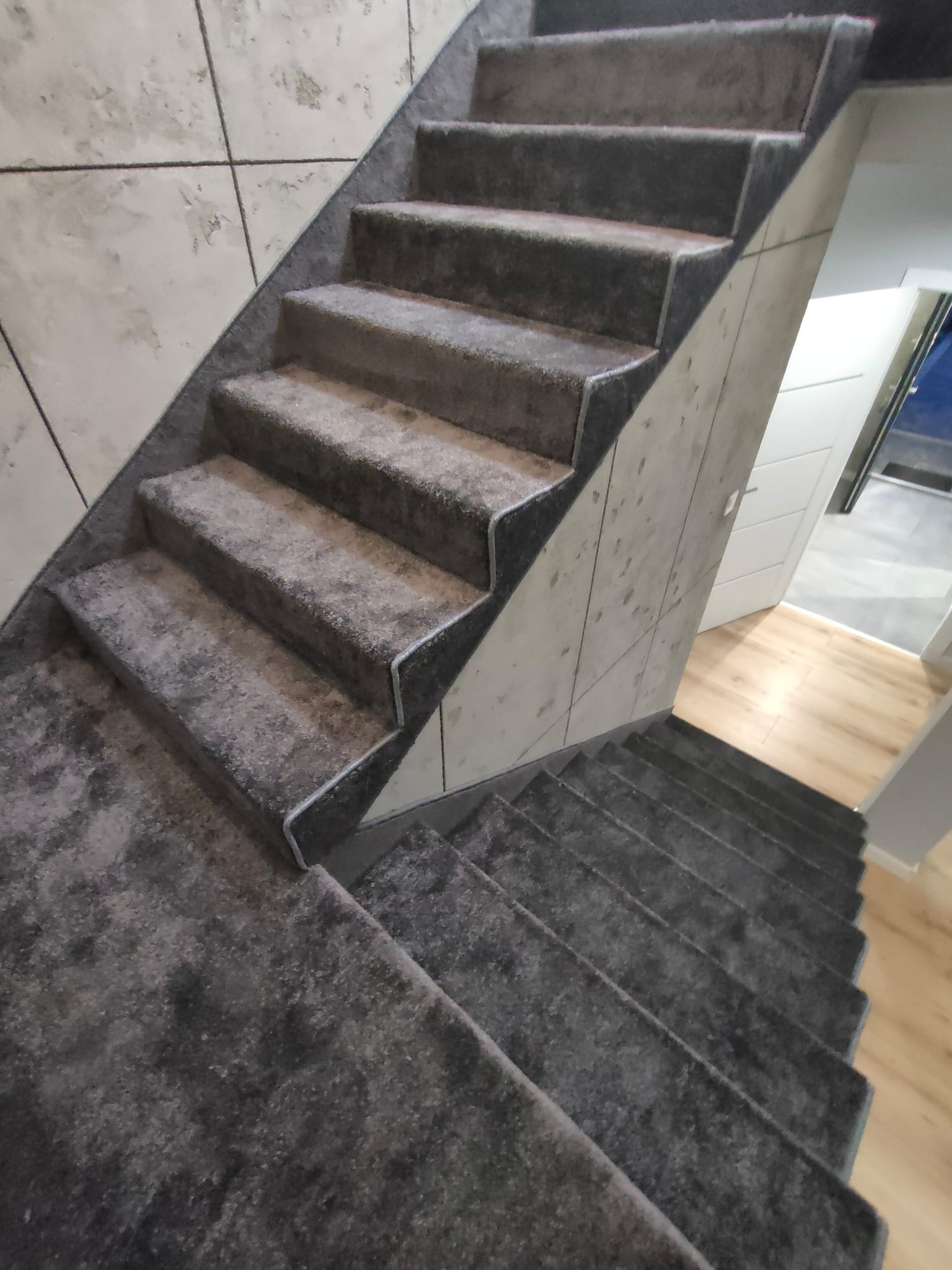 Schody wyłożone szarą wykładziną dywanową z ciemnym obramowaniem, widok z góry. W tle jasna ściana z efektem betonu i fragment podłogi z paneli.