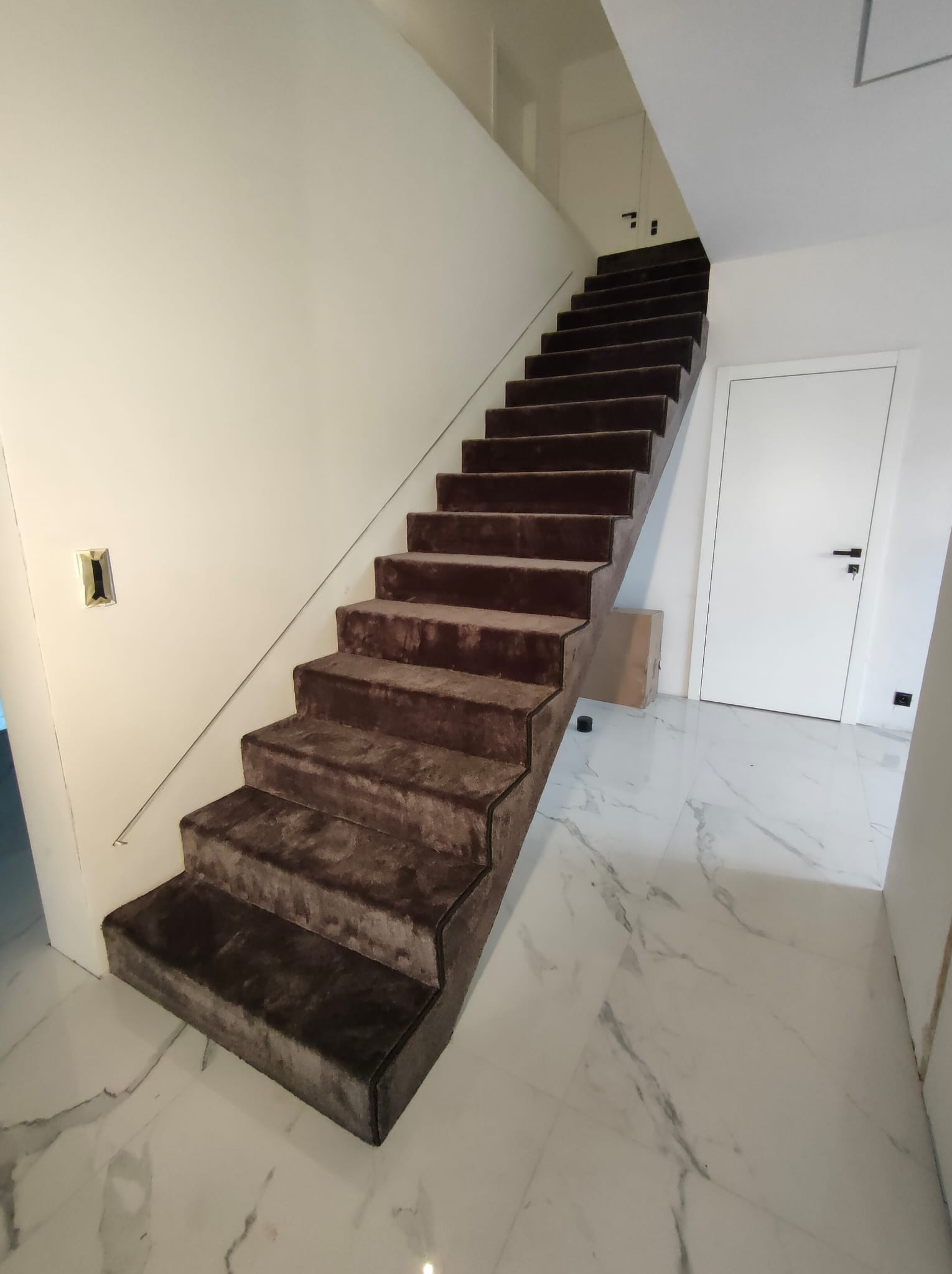 Schody wyłożone brązową wykładziną dywanową na białych marmurowych płytkach. Nowoczesne wnętrze z minimalistyczną balustradą i białymi ścianami.