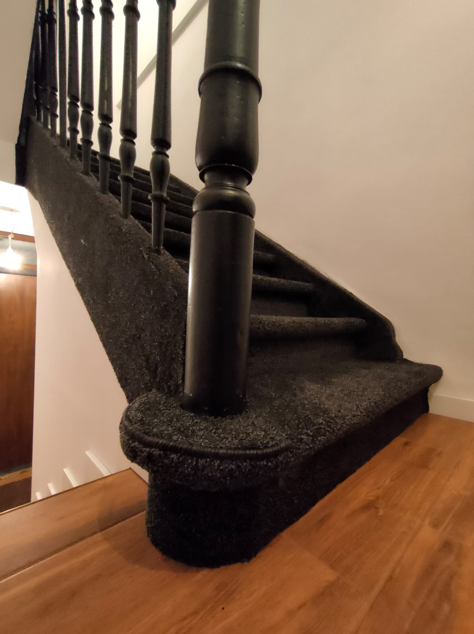 Schody wyłożone ciemną, teksturowaną wykładziną, z czarnymi drewnianymi balustradami. Widoczny fragment drewnianej podłogi na dole schodów.