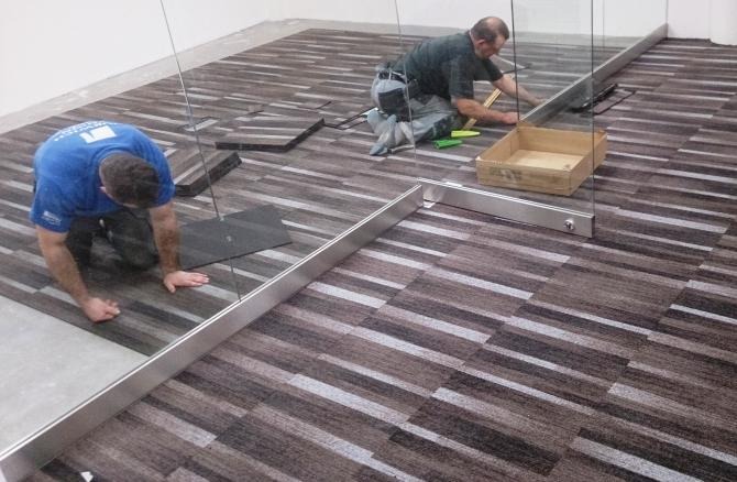 Układanie brązowo-szarej wykładziny dywanowej w płytkach. Dwóch monterów pracuje w pomieszczeniu biurowym, widoczne narzędzia pracy i fragment szklanej ścianki działowej.