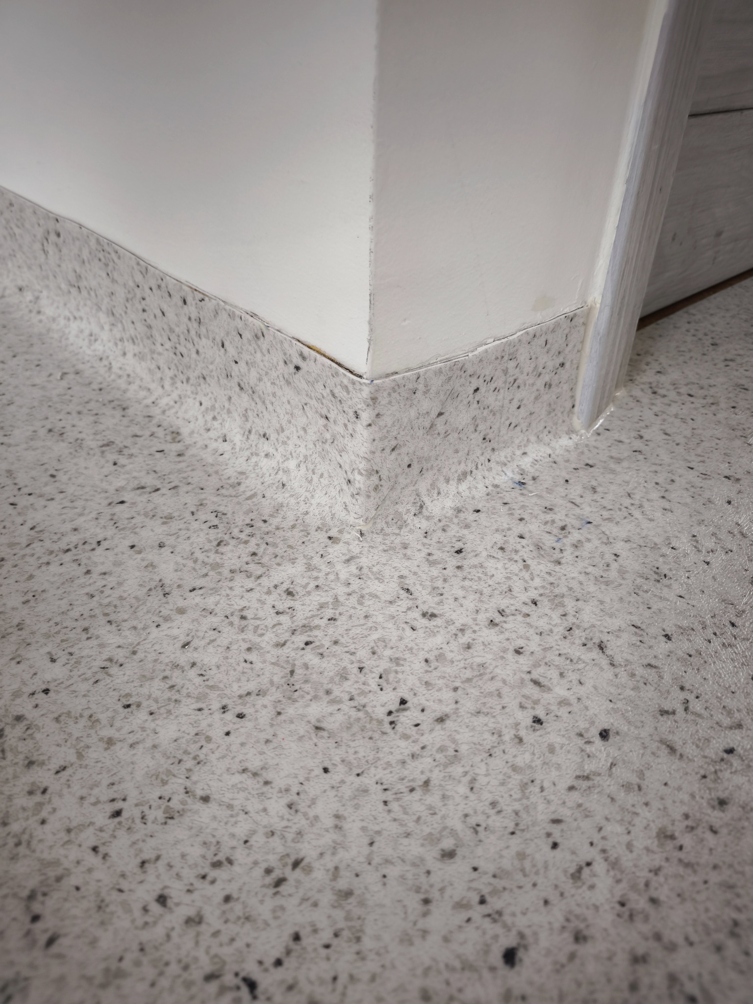 Szara wykładzina podłogowa z drobnymi, ciemnymi plamkami, idealnie dopasowana do listew przypodłogowych i ściany w rogu pomieszczenia. Precyzyjne wykończenie.