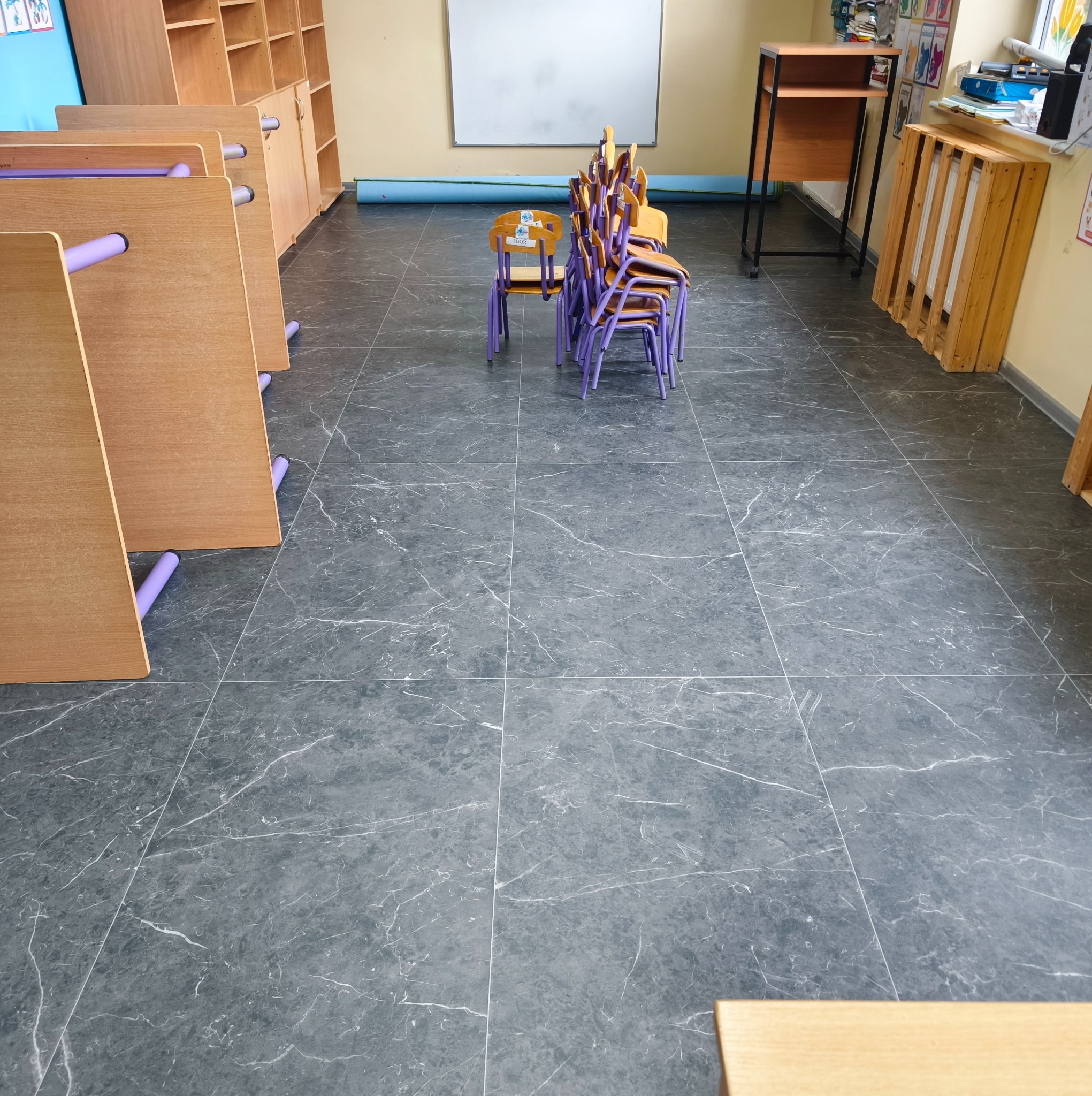 Szara, marmurkowa podłoga w sali przedszkolnej z ułożonymi krzesłami i stolikami. Widok na całą powierzchnię podłogi, ukazujący wzór i wykończenie.