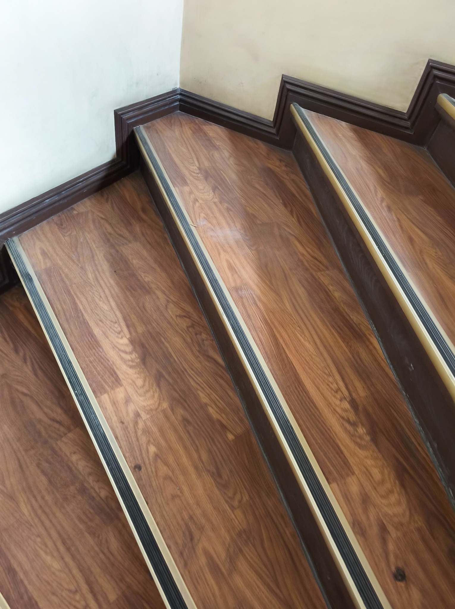 Schody wykończone panelami drewnopodobnymi z listwami antypoślizgowymi, widoczne z góry. Ciemnobrązowe wykończenie przy ścianie. Detal wnętrza.