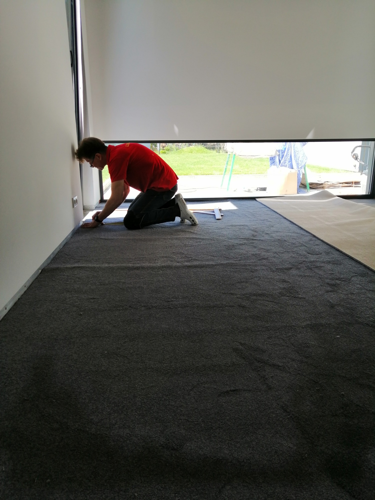 Pracownik w czerwonej koszulce na kolanach przy ścianie, docina szarą wykładzinę w jasnym pomieszczeniu z dużym oknem i roletą.