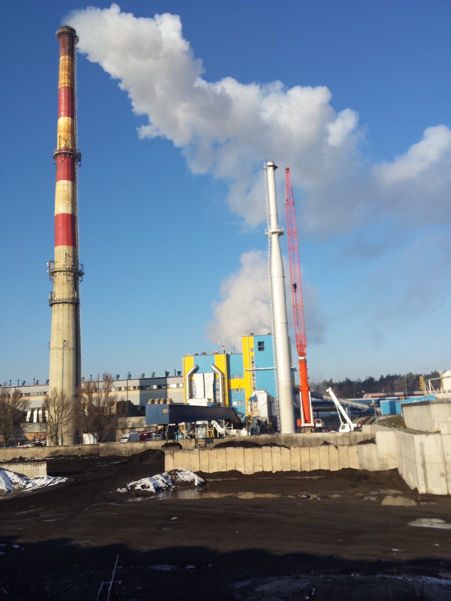 Wysoki, ceglano-biały komin z widocznymi instalacjami, obok komin w kolorze srebrnym z czerwoną dźwignią na tle błękitnego nieba z chmurami i fragmentem zabudowań przemysłowych.