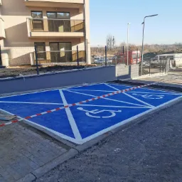 Oznakowane na niebiesko miejsca parkingowe dla osób niepełnosprawnych przed nowym budynkiem mieszkalnym, zabezpieczone taśmą ostrzegawczą.
