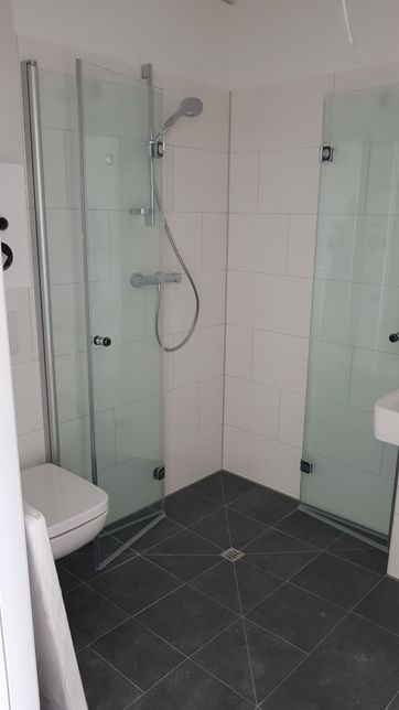 Nowoczesna łazienka z kabiną prysznicową ze szklanymi drzwiami na zawiasach, białe płytki ścienne i ciemnoszara podłoga z odpływem liniowym.