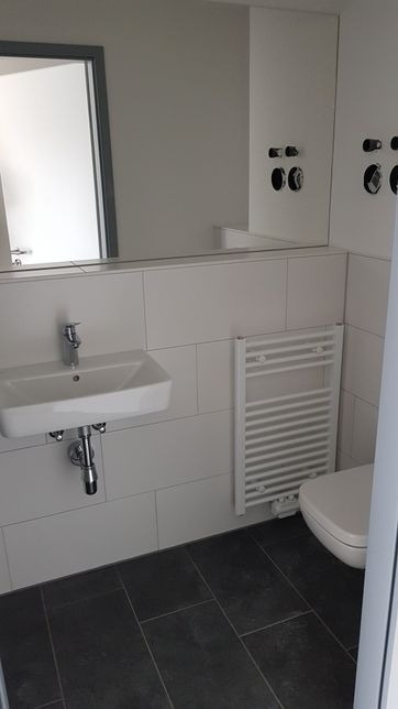 Nowoczesna łazienka z białymi płytkami imitującymi cegłę, wiszącą umywalką, grzejnikiem drabinkowym i ciemną podłogą z płytek w formacie prostokątnym; widoczne przygotowane punkty elektryczne.