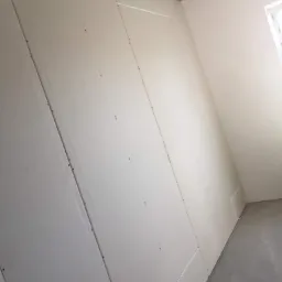 Ściana w trakcie montażu płyt karton-gips z widocznymi łączeniami i wkrętami, surowy betonowy sufit z wystającymi przewodami elektrycznymi, fragment okna w prawym górnym rogu.