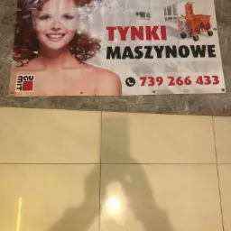 Baner reklamowy z uśmiechniętą kobietą w masce, maszyną do tynkowania i numerem telefonu na tle wnętrza budynku