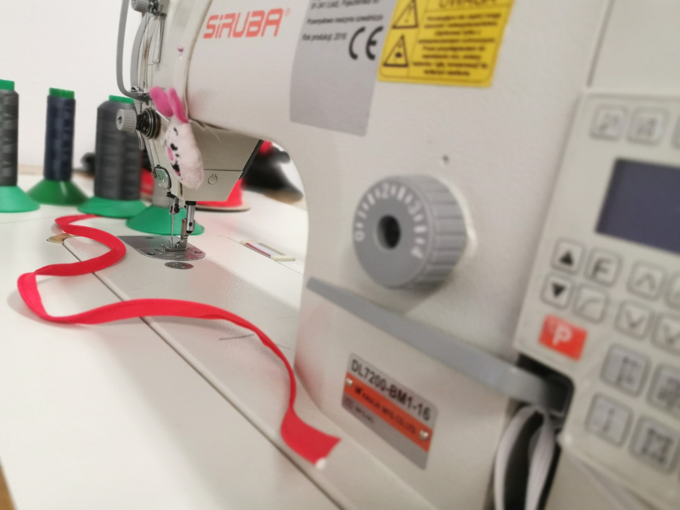 Profesjonalna maszyna do szycia marki Siruba, z czerwoną wstążką pod stopką i ozdobą w kształcie królika przy igle, w tle widoczne szpulki nici.