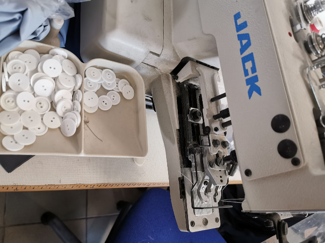 Zbliżenie na maszynę szwalniczą marki Jack z pojemnikiem pełnym białych guzików dwudziurkowych, widoczne elementy mechaniczne maszyny.