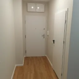 Wnętrze przedpokoju po remoncie z panelami podłogowymi imitującymi drewno, białe ściany, białe drzwi wejściowe i wewnętrzne z dekoracyjnym wzorem w jodełkę oraz biała szafka elektryczna pod sufitem.