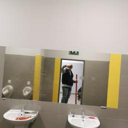 Ale Chata - Duże lustro w publicznej toalecie z szarymi płytkami i żółtymi pasami, odbijające mężczyznę z poziomicą i częściowo zakryte plamami, z umywalkami i pisuarami widocznymi w pomieszczeniu.