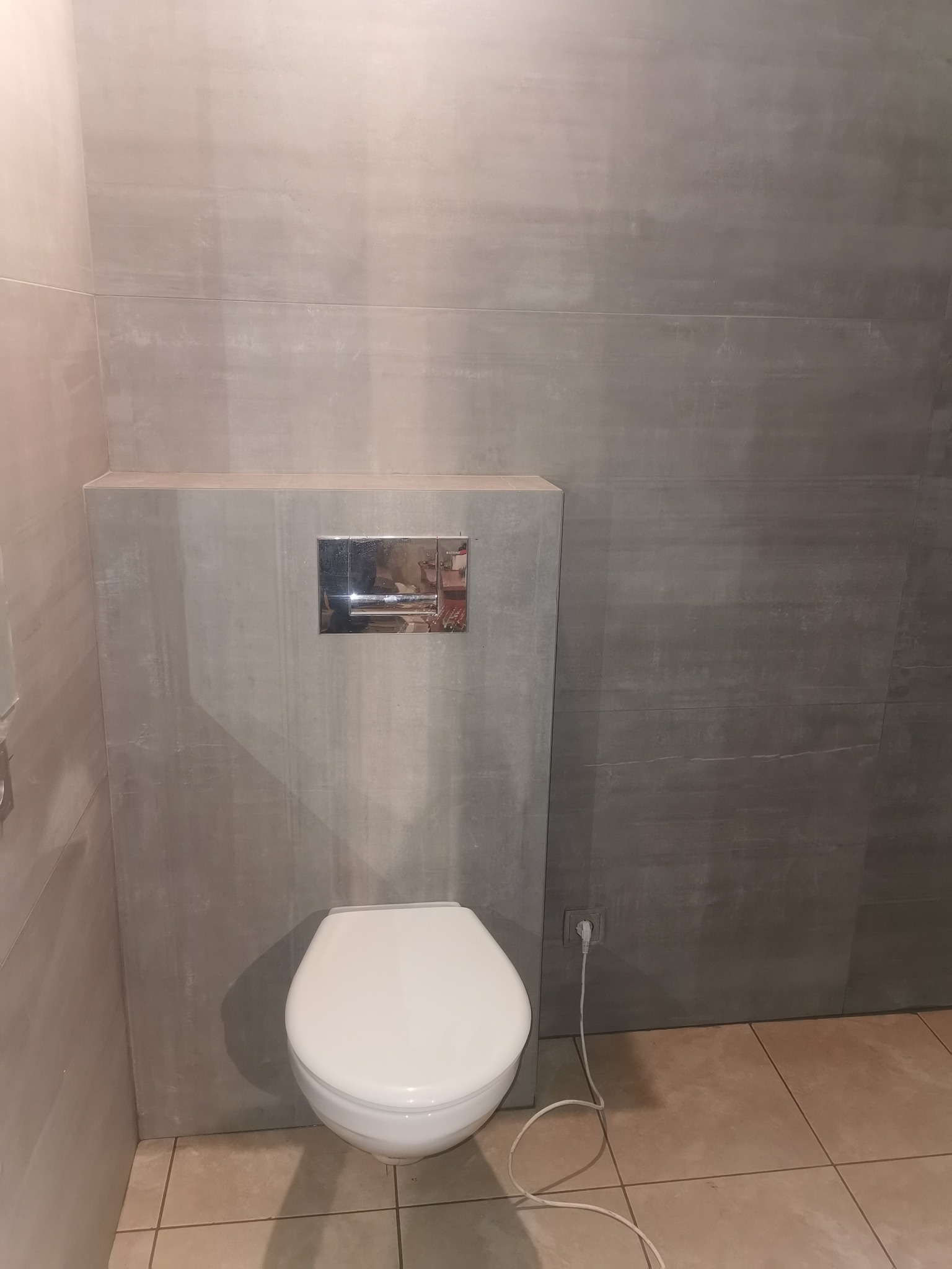 Nowoczesna toaleta podwieszana z szarymi płytkami imitującymi beton na ścianach, widoczny chromowany przycisk spłuczki i gniazdko elektryczne z białą wtyczką po prawej stronie.