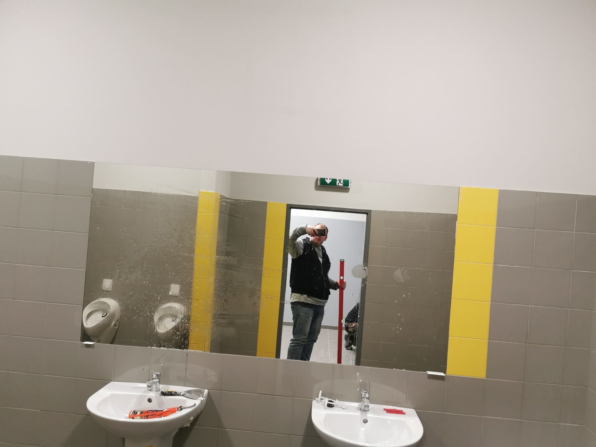 Duże lustro w publicznej toalecie z szarymi płytkami i żółtymi pasami, odbijające mężczyznę z poziomicą i częściowo zakryte plamami, z umywalkami i pisuarami widocznymi w pomieszczeniu.