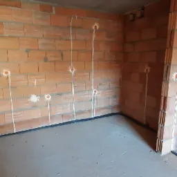 Surowy stan wnętrza z widocznymi przewodami elektrycznymi wychodzącymi ze ścian z cegły, przygotowane do dalszych prac wykończeniowych.