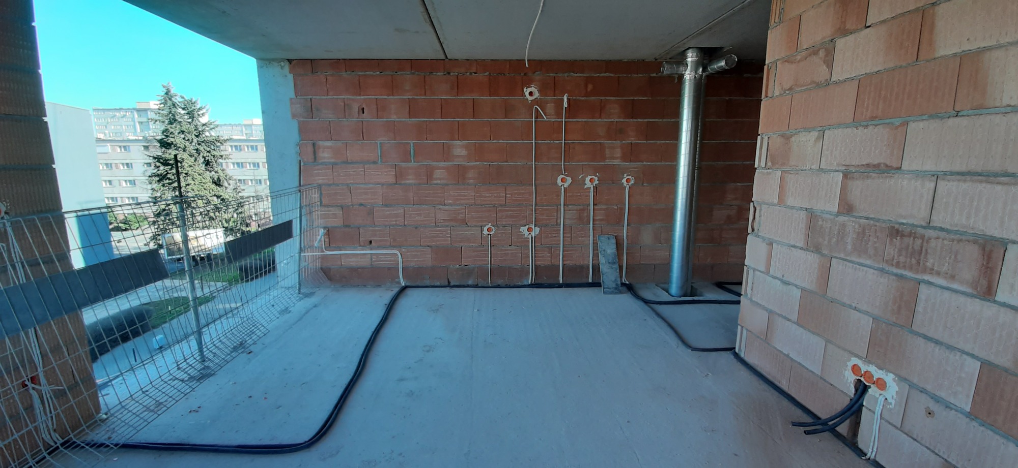 Surowy stan balkonu w bloku z widocznymi przewodami elektrycznymi wychodzącymi ze ściany i rurą wentylacyjną. W tle blok mieszkalny i drzewo.