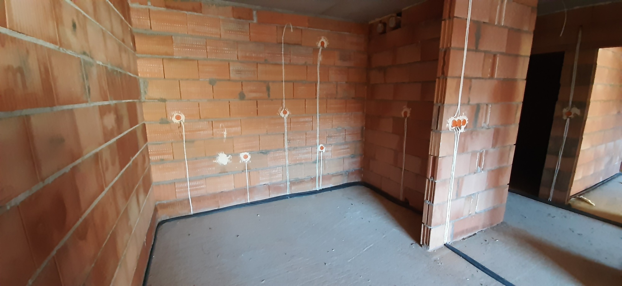 Surowy stan wnętrza z widocznymi przewodami elektrycznymi wychodzącymi ze ścian z cegły, przygotowane do dalszych prac wykończeniowych.