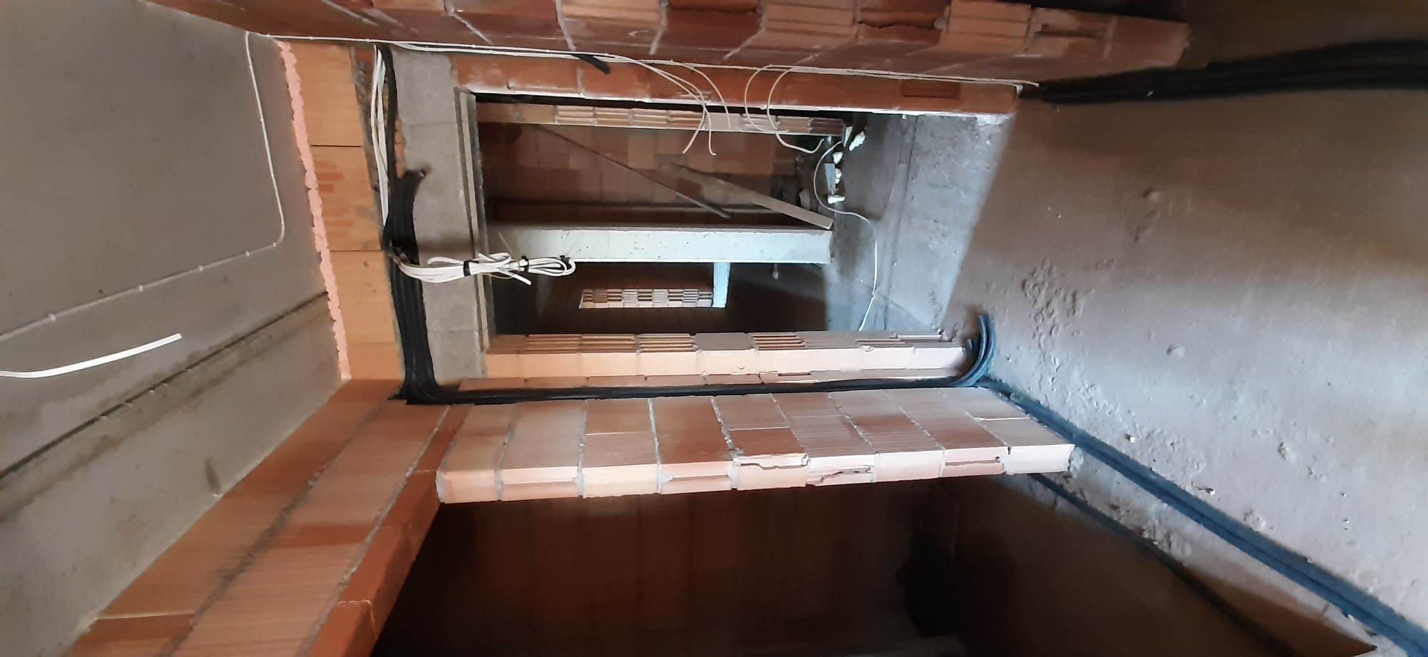 Kable elektryczne prowadzone wzdłuż muru z cegły, widok z góry na pionowy szyb instalacyjny w surowym stanie.