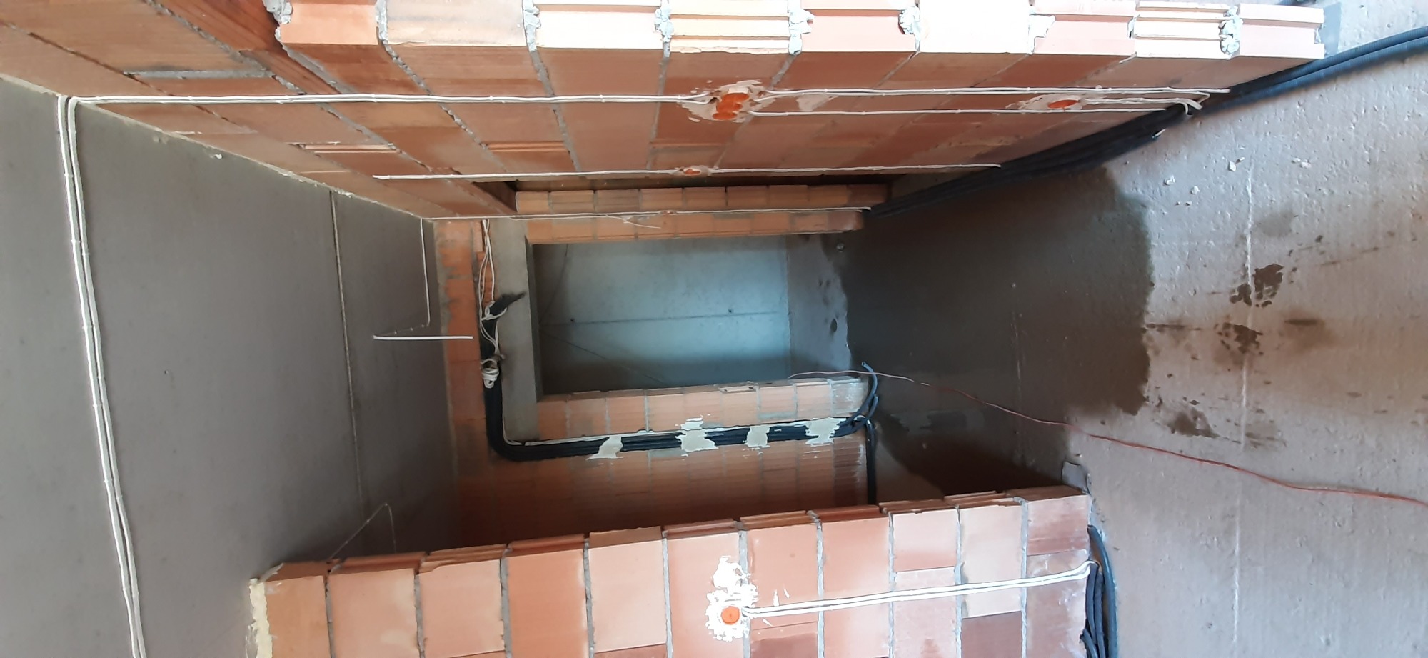 Wnętrze szybu wentylacyjnego w trakcie budowy, widoczne ceglane ściany z rozprowadzoną instalacją elektryczną w białych peszlach, surowy beton na ścianach.