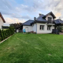 Nowoczesny dom z zadbanym trawnikiem i tujami wzdłuż granicy działki. Widoczna huśtawka i zadaszony taras. Ujęcie z szerokiego kąta.