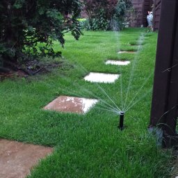 RAUT ROBERT WOJDAK - Automatyczny system nawadniania trawnika w ogrodzie. Zraszacze rozpylają wodę na soczystą trawę, obok ułożone są kwadratowe płyty chodnikowe.