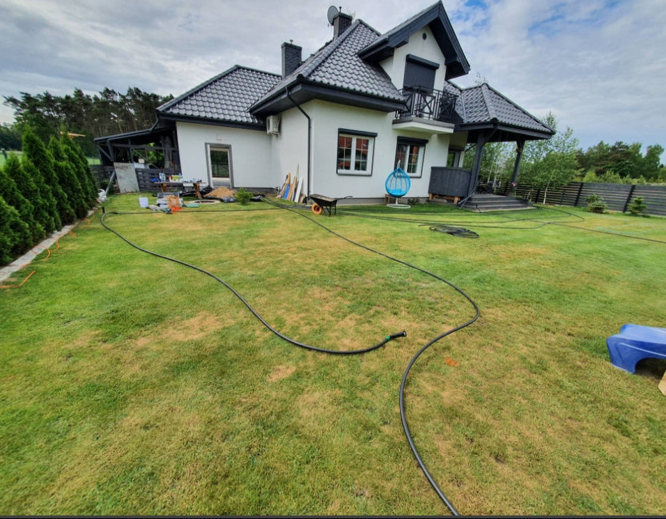 Rozłożony system nawadniania na trawniku przed domem z ciemną dachówką. W tle widoczne drzewa i krzewy. Węże nawadniające na trawie.