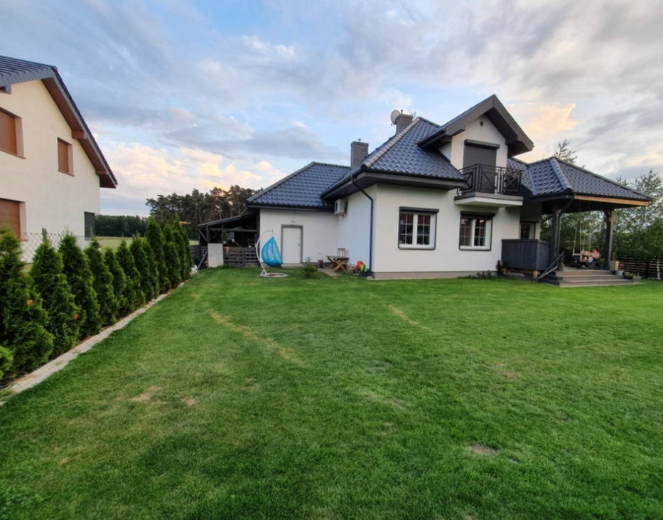 Nowoczesny dom z zadbanym trawnikiem i tujami wzdłuż granicy działki. Widoczna huśtawka i zadaszony taras. Ujęcie z szerokiego kąta.