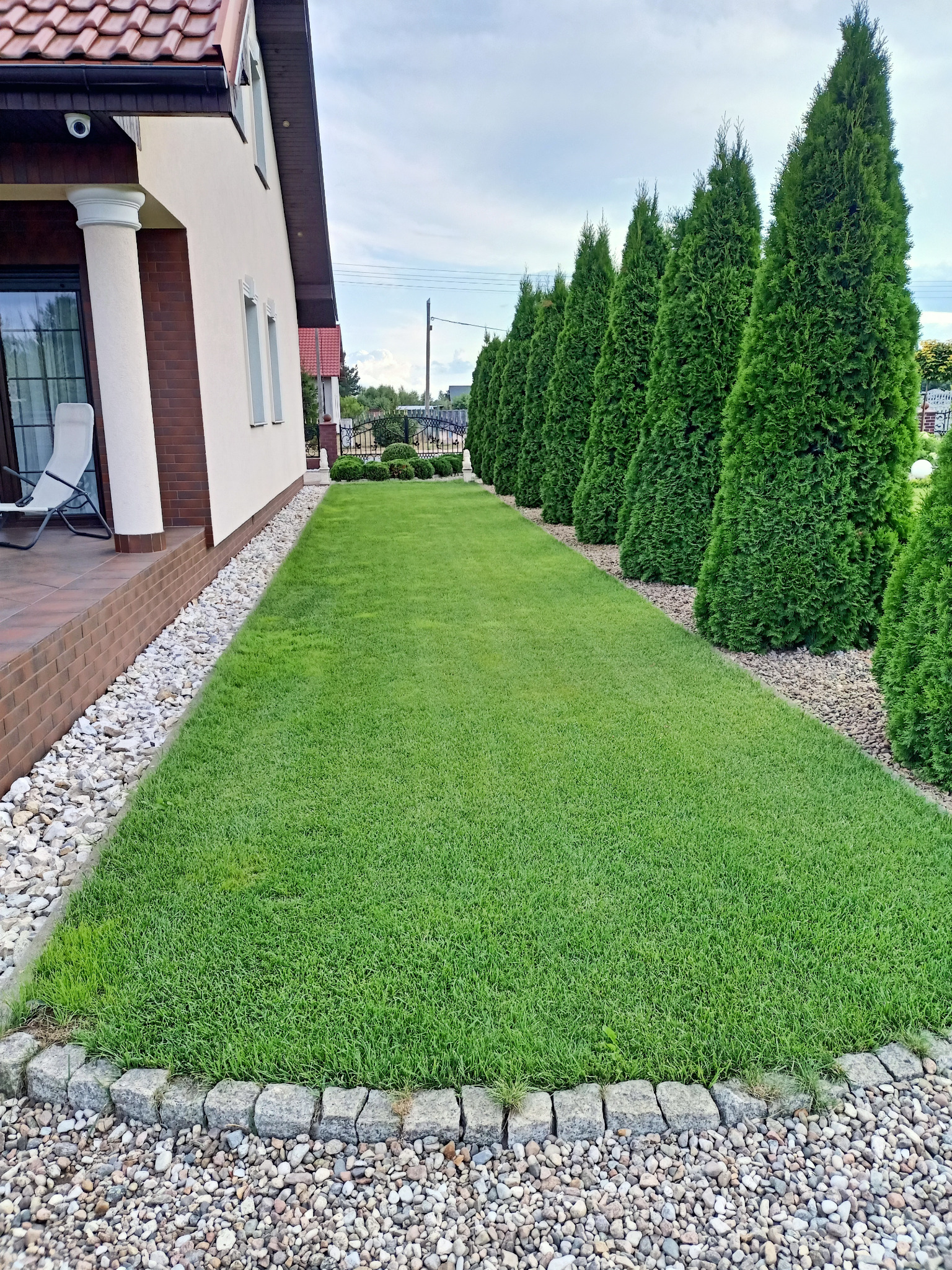 Zadbany trawnik z równo przyciętą trawą, obrzeżem z kamienia i szpalerem tuj wzdłuż ogrodzenia. Widok na elewację domu z oknem i kolumnami.