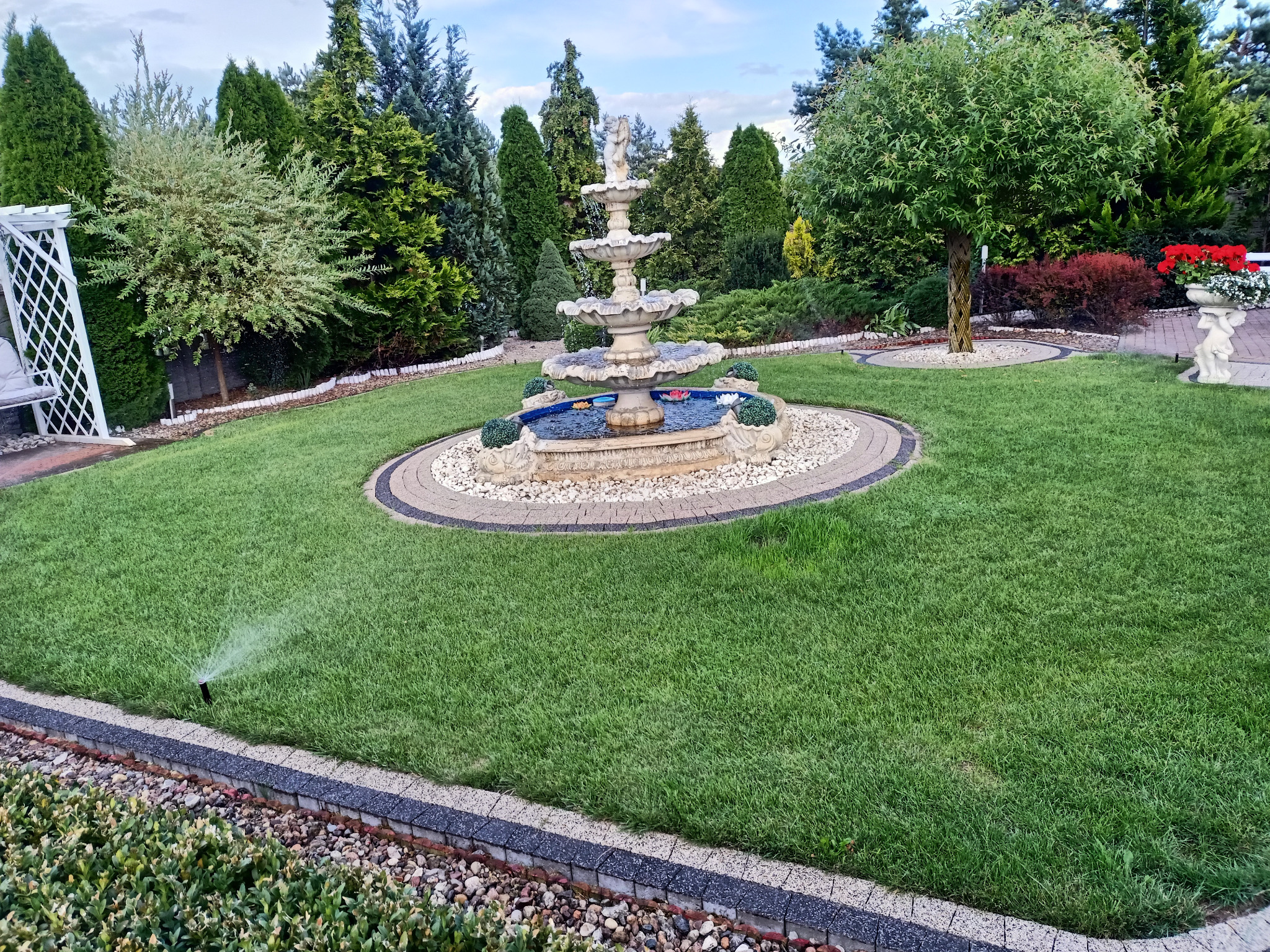 Utrzymany trawnik z fontanną i systemem nawadniania. W tle różnorodne drzewa i krzewy. Woda tryska z dyszy nawadniającej.