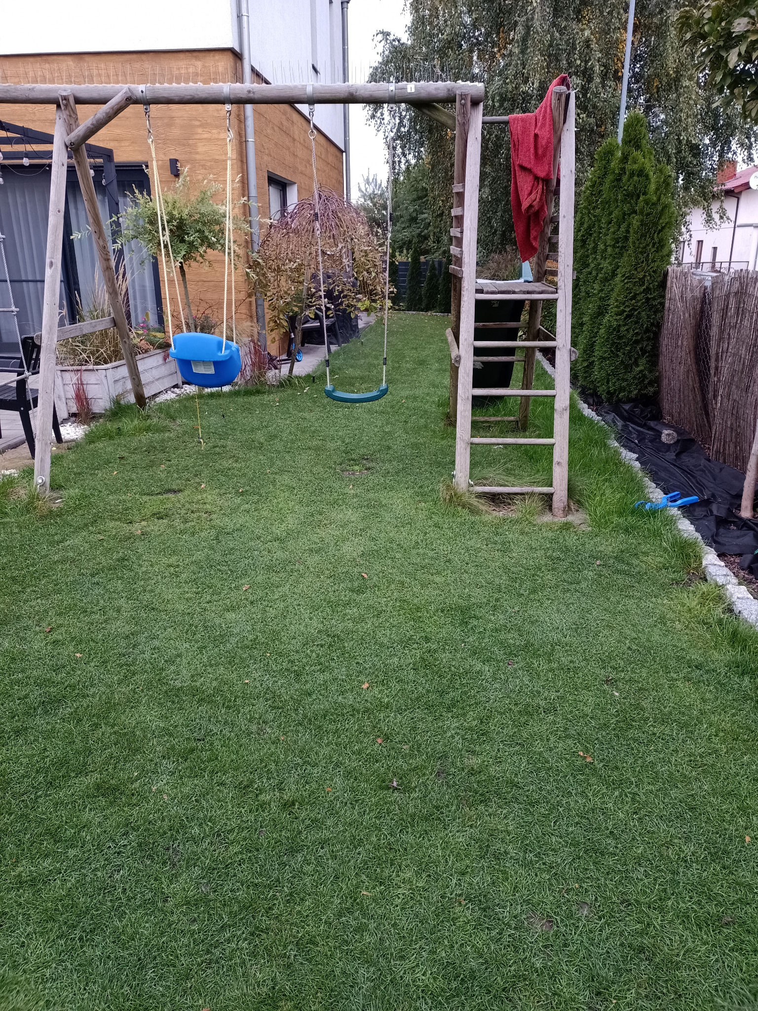 Zadbany trawnik z huśtawkami i elementami placu zabaw w ogrodzie, widok na dom z drewnianą elewacją i roślinność.