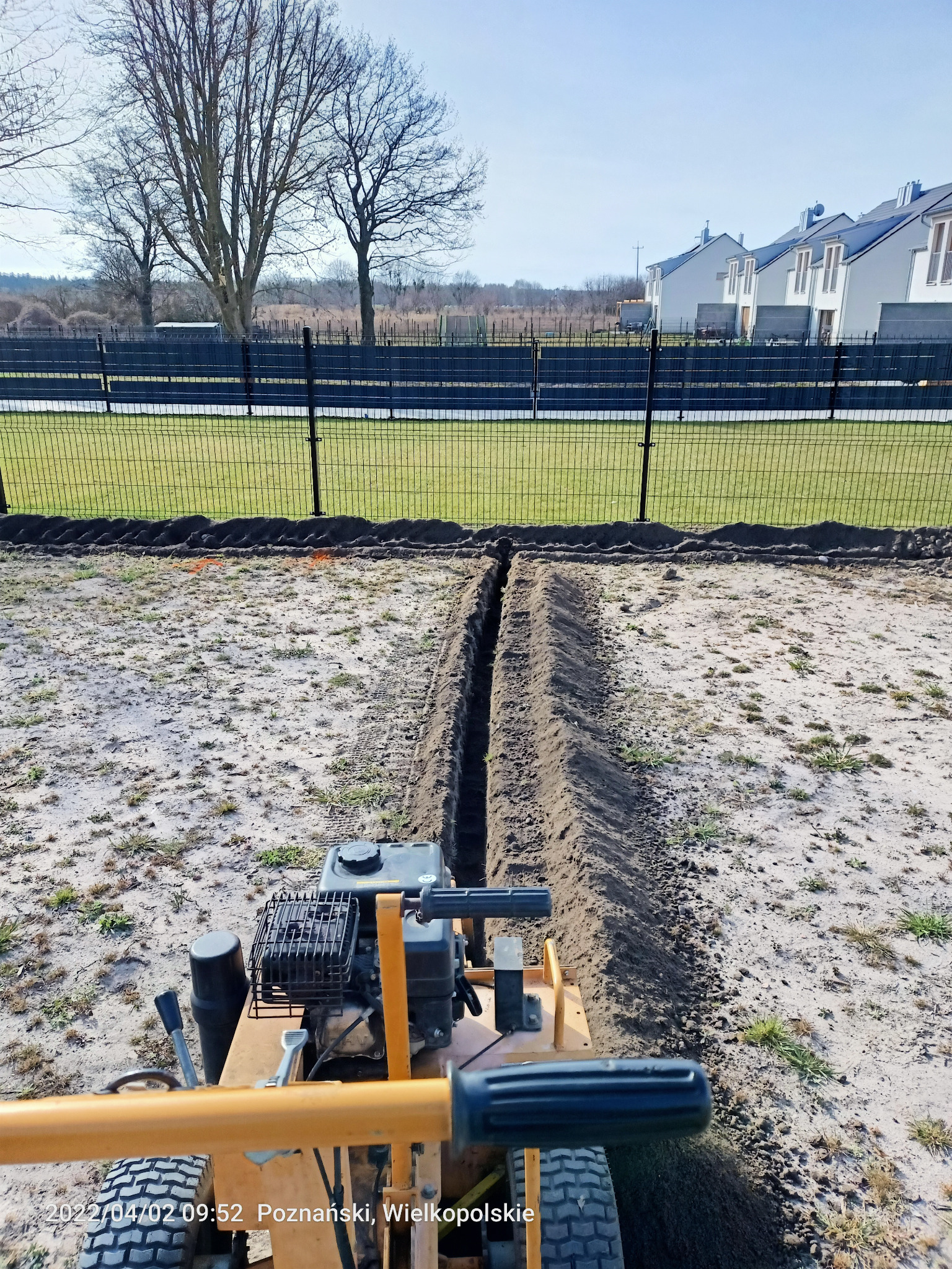 Wykonanie wykopu pod system nawadniania w ogrodzie przy użyciu specjalistycznej maszyny. W tle ogrodzenie i zabudowa jednorodzinna.