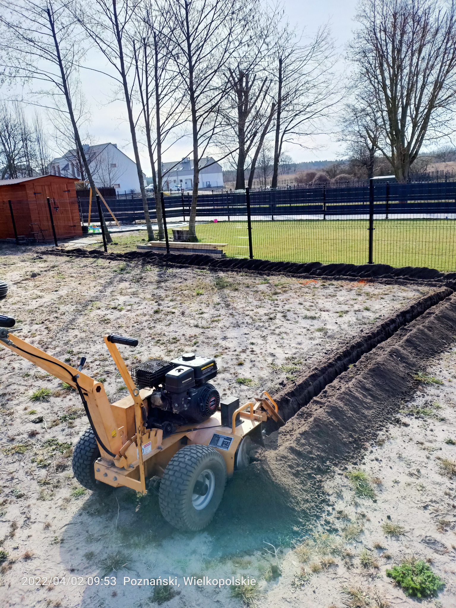 Żółta glebogryzarka wykopuje rów w ziemi na działce, przygotowując teren pod system nawadniania. Widoczny czarny płot i budynek w tle.