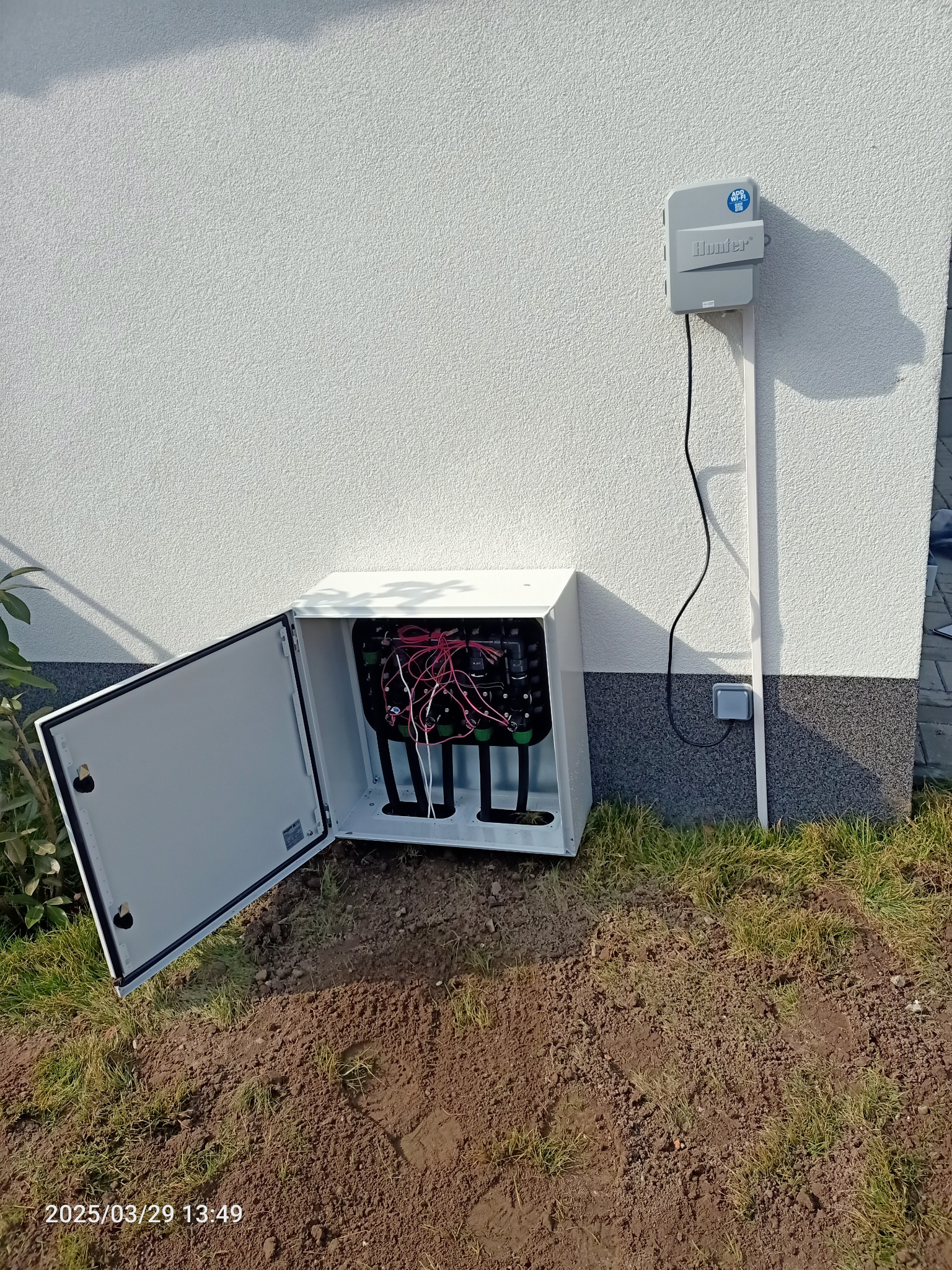 Otwarta skrzynka sterownicza systemu nawadniania Hunter, zamontowana na elewacji domu, z widocznymi przewodami i elementami instalacji.