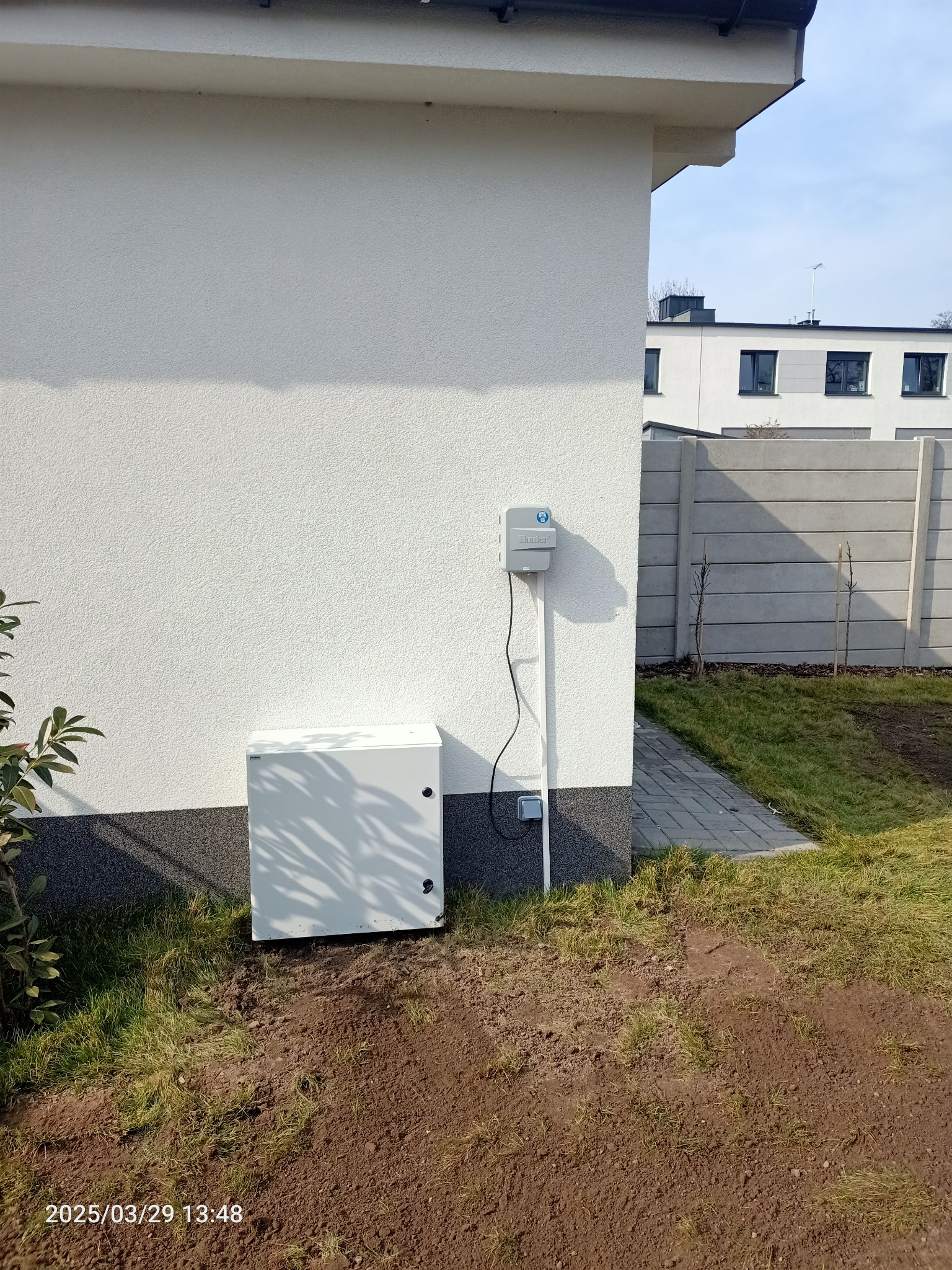 Panel sterowania systemem nawadniania Hunter, zamontowany na elewacji domu w Obornikach. Widoczna skrzynka zasilająca i przewody. Trawa i świeżo przekopana ziemia.