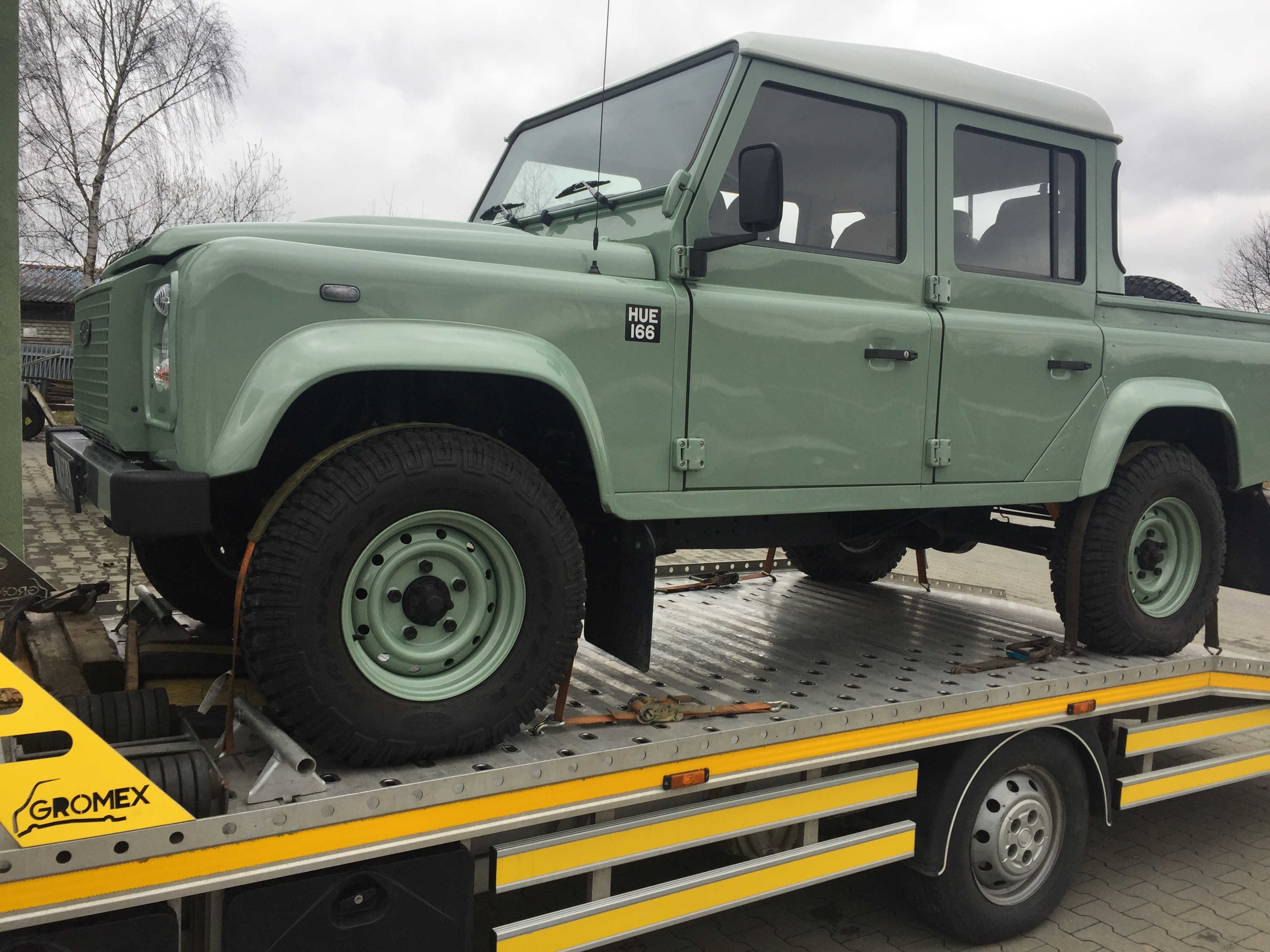 Jasnozielony Land Rover Defender na żółto-srebrnej platformie lawety, zabezpieczony pasami transportowymi, z widocznym logo firmy Gromex.