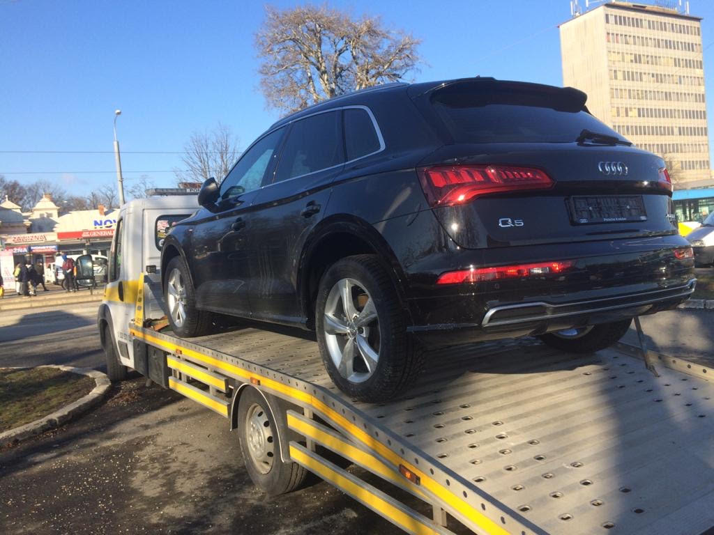 Czarny samochód osobowy Audi Q5 na platformie niskopodwoziowej z żółtymi elementami, w tle budynek i drzewo bez liści w słoneczny dzień.