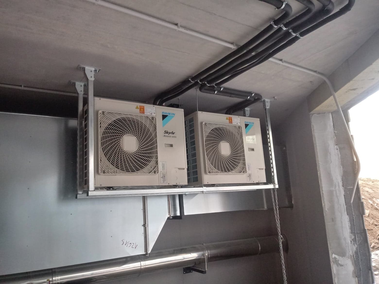Dwa agregaty zewnętrzne Daikin SkyAir zamontowane pod betonowym stropem, widoczne rury instalacyjne i elementy mocujące, ślady pisma na metalowej obudowie wentylacji.