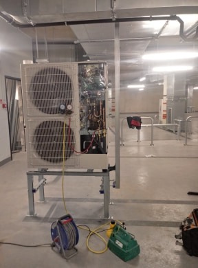 Dwuczęściowa jednostka zewnętrzna z otwartą obudową, widoczne miedziane rurki i okablowanie, zamontowana na metalowej konstrukcji w pomieszczeniu technicznym.