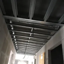 Metalowa konstrukcja sufitu podwieszanego w trakcie montażu, widoczne profile i przewody wentylacyjne, ściany w stanie surowym z widocznymi łączeniami płyt gipsowo-kartonowych.