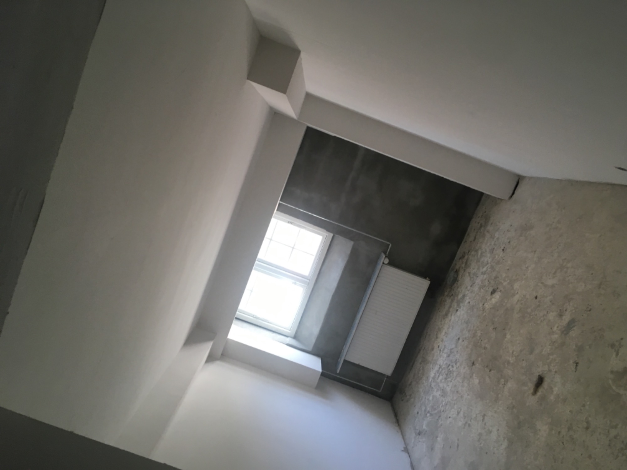Wnętrze w trakcie remontu, widok z dołu na okno, grzejnik i surową ścianę, częściowo pomalowane ściany na biało, surowy beton na podłodze.