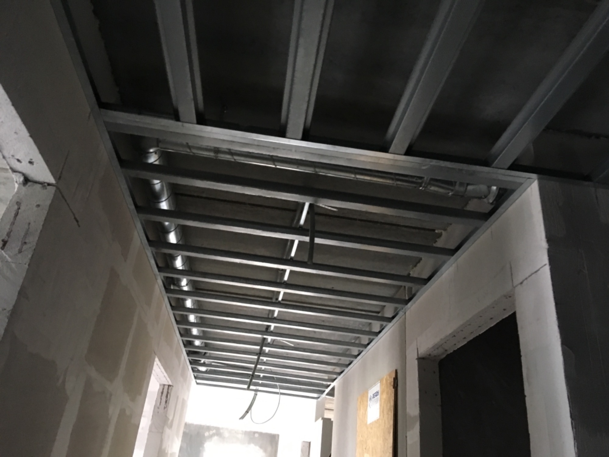 Metalowa konstrukcja sufitu podwieszanego w trakcie montażu, widoczne profile i przewody wentylacyjne, ściany w stanie surowym z widocznymi łączeniami płyt gipsowo-kartonowych.