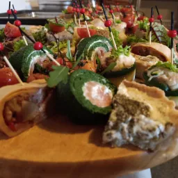 Deska z różnorodnymi przekąskami: zielone roladki ze szpinaku i łososia, mini tartaletki z farszem, pomidorki koktajlowe na wykałaczkach, koreczki z warzyw i pieczywa, dekorowane zieleniną.