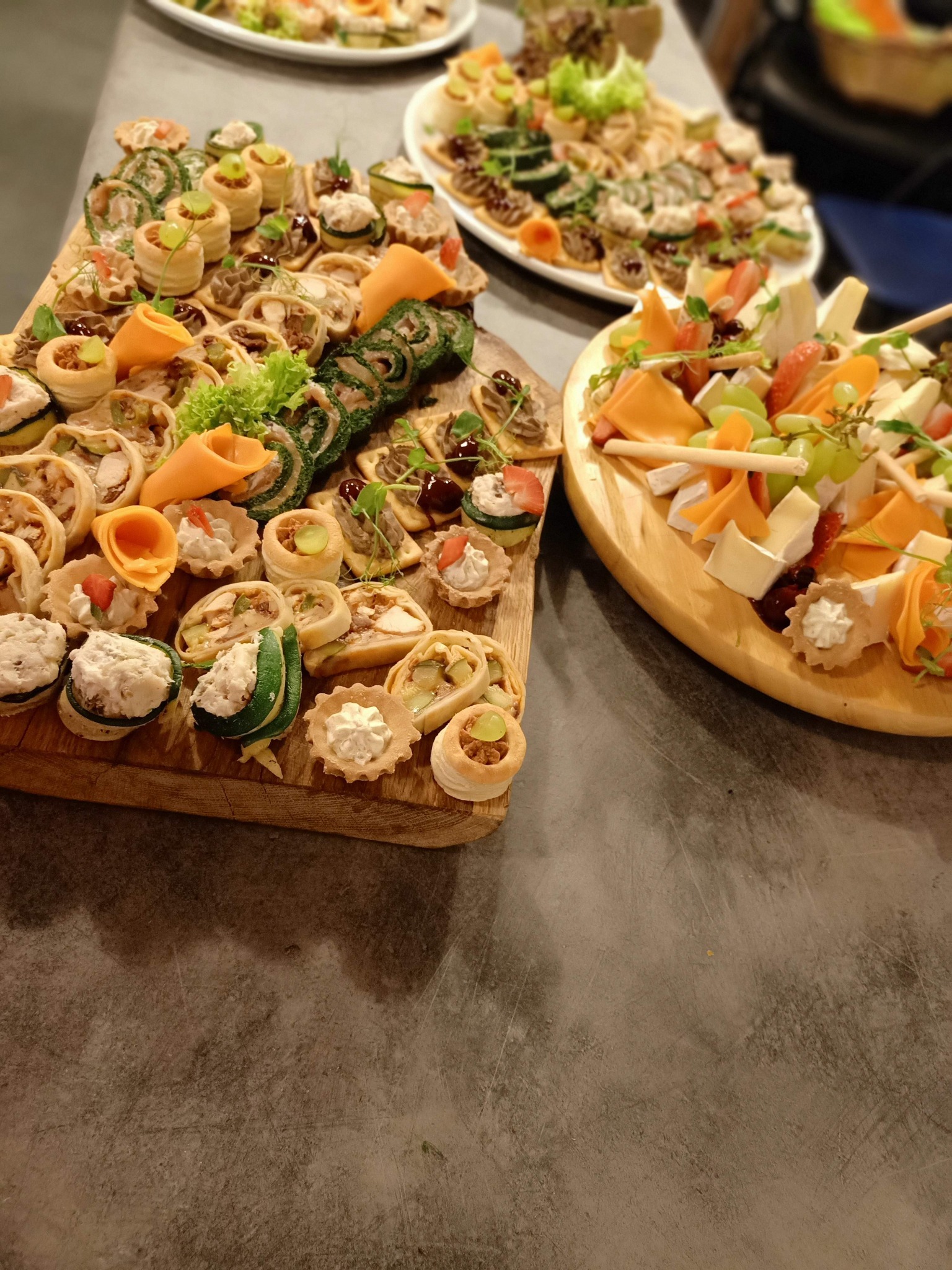 Drewniane deski i biały talerz z różnorodnymi, apetycznymi przekąskami: tartaletki z kremem, roladki z warzywami, koreczki z serem i owocami, ułożone na stole w neutralnych barwach.