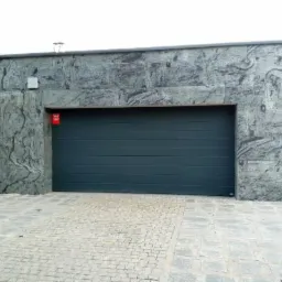 Nowoczesny garaż z elewacją z szarego kamienia o unikalnym wzorze, dopasowana kolorystycznie brama garażowa, kostka brukowa na podjeździe.