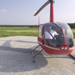 Szydzisz - Czerwony helikopter Robinson R44 z pilotem w kokpicie, stojący na betonowym lądowisku z widocznym cieniem śmigła, w tle zielone pole i las.