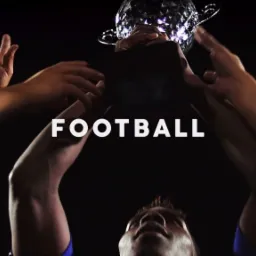 Ujęcie od dołu: Ręce unoszą błyszczący puchar nad głową, ciemne tło, widoczna twarz osoby w dolnej części kadru.