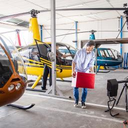 Szydzisz - Ekipa filmowa w hangarze podczas nagrywania ujęć z helikopterami w tle, operator kamery na podwyższeniu, oświetlenie i rekwizyty rozstawione.